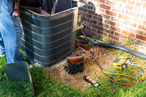 HVAC technician servicing an outdoor air conditioning condenser unit beside a brick home with tools and gauges on the lawn
